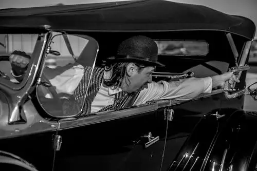 Free Dramatic black and white photo of a vintage 1930s-style gangster aiming a pistol from a classic car. Stock Photo